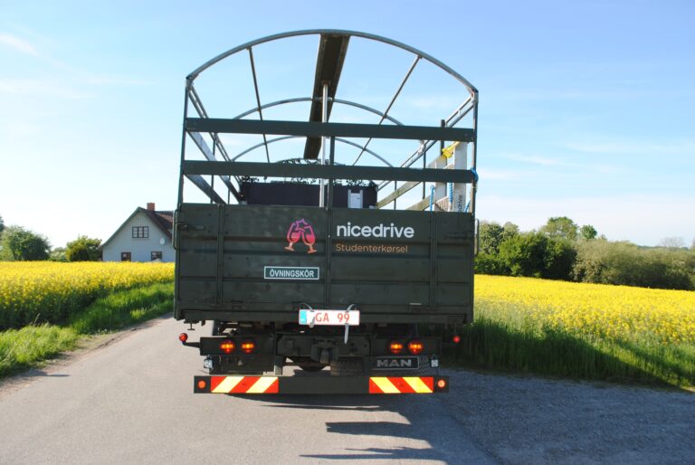 Bagsiden af Nicedrive militærlastbil til studenterkørsel, med logo og åbent lad, klar til festlig transport af studenter gennem naturskønt landskab